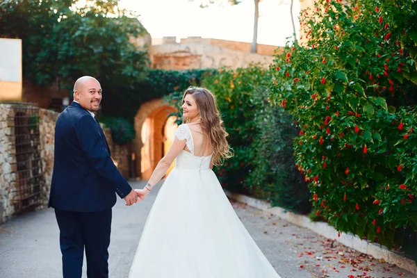 The newlyweds walk and smile, at the entrance to the castle against the background of a stone wall and palm trees.