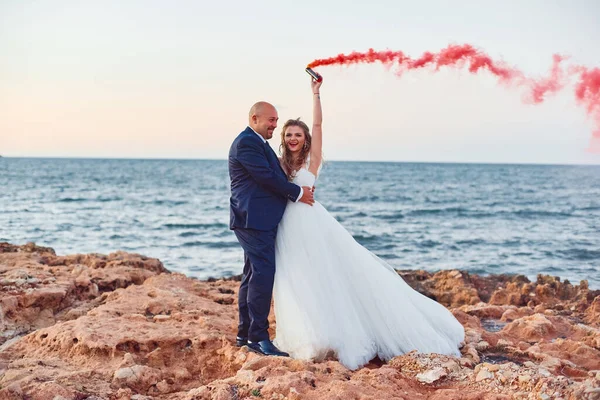花嫁は海岸や岩で赤い煙を上げて笑顔で新郎を抱きしめる — ストック写真