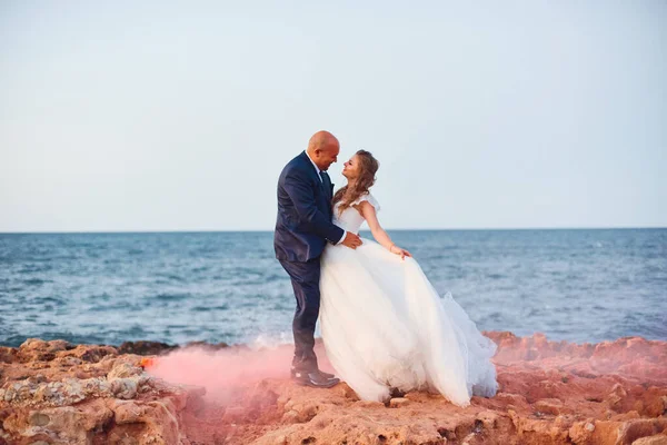 花嫁は海岸や岩の赤い煙を背景に笑顔で新郎を抱きしめる — ストック写真