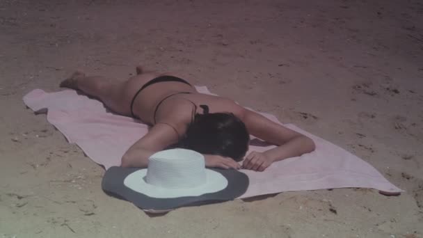 Брюнетка в черном купальнике и шляпе загорает на пляже у моря в летний солнечный день — стоковое видео