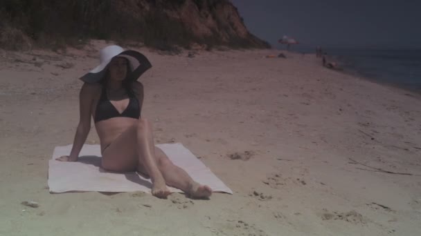 Chica morena en traje de baño negro y sombrero tomando el sol en la playa junto al mar en verano día soleado — Vídeos de Stock