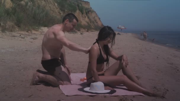 Guy anoints sunscreen on the back of girl while relaxing on the sea beach in summer — Stok Video