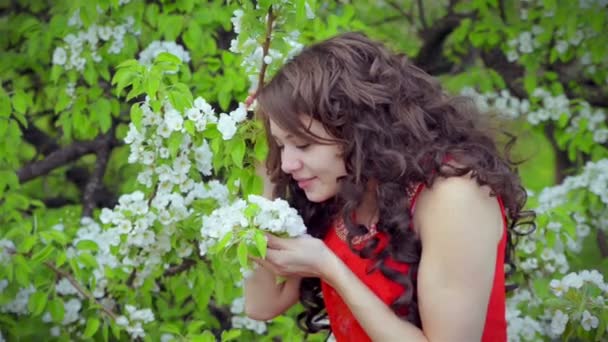 Glad flicka i röd klänning att lukta blommor i vårträdgård och ler. — Stockvideo