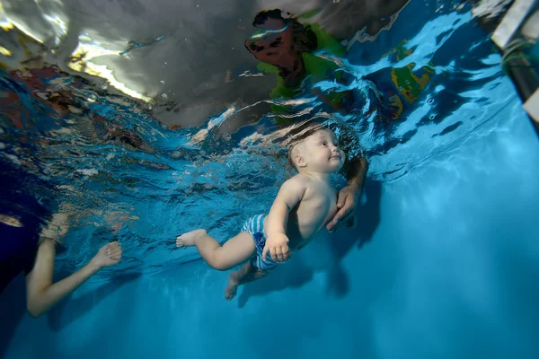 Feliz bebé entrena y nada bajo el agua en la piscina sobre un fondo azul, y el padre lo atrapa — Foto de Stock