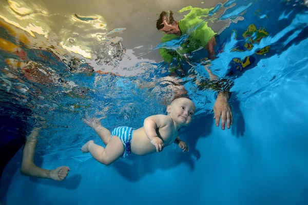 Feliz niño entrena y nada bajo el agua en la piscina sobre un fondo azul, y mamá y papá lo ayudan — Foto de Stock