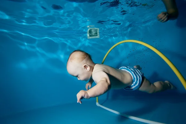 Le bébé est formé pour nager à travers un cerceau sous l'eau dans la piscine sur un fond bleu. Gros plan. Orientation horizontale — Photo