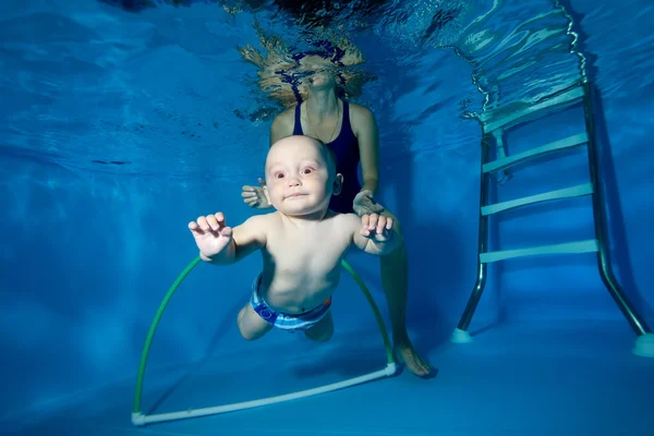 Mamá enseña y entrena al bebé a nadar a través de un aro bajo el agua en la piscina sobre un fondo azul. Primer plano. Retrato. Orientación horizontal . — Foto de Stock