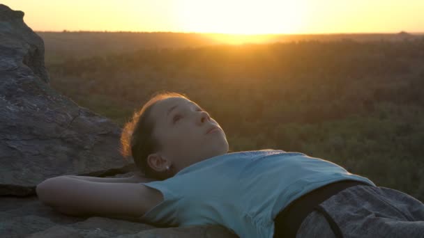 Uma jovem adolescente está deitada, olhando para o céu e sonhando em um topo da montanha com um fundo de pôr do sol. Retrato feminino. Close-up. 4K. — Vídeo de Stock