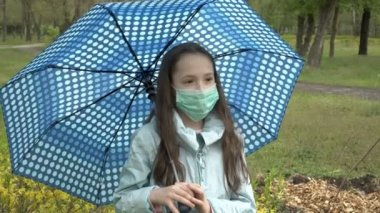 Yüzünde tıbbi maske takmış, kasvetli bir sonbahar gününde bir şehir parkında şemsiye altında poz veren üzgün bir genç kızın portresi. Yakın plan. Kavram. 4K.