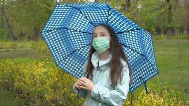 Una bella adolescente con una maschera medica sul viso si trova sotto un ombrello in un parco cittadino in una cupa giornata autunnale. Primo piano. Ritratto femminile. Concetto. 4K. — Video Stock