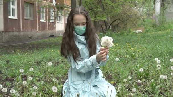 Молодая девушка-подросток в медицинской маске на лице собирает букет цветов из белых одуванчиков на заднем дворе дома. Закрывай. Портрет женщины. 4K — стоковое видео