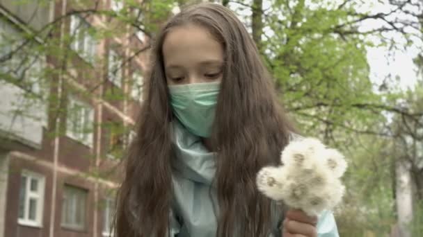 Retrato de una pequeña adolescente con una máscara médica en la cara con un ramo de dientes de león blancos en la mano. Recoge flores en el patio trasero. Primer plano. Vista de abajo. 4K. — Vídeo de stock
