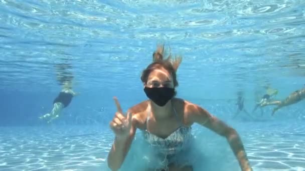 A beautiful teenage girl wearing a black face mask poses for the camera underwater in a pool in a shiny dress. She looks at the camera and waves a warning hand. Closeup. Female portrait. Slow motion — Stock Video