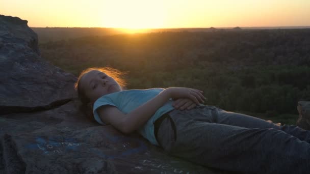 Uma menina feliz jaz e descansa ao pôr do sol no topo de uma montanha acima de um vale verde. Ela sorri e olha atentamente para o céu. Conceito. Fecha. Retrato. 4K. — Vídeo de Stock