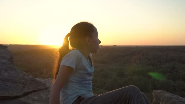 Beautiful young teen girl sitting on top of a mountain with sunset background. Below it you can see a mountain valley overgrown with forest. Female portrait. Close-up. 4K. — Stock Video