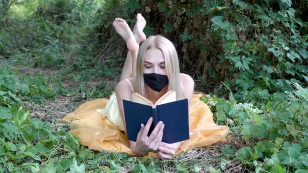 Linda rubia con una máscara protectora está mintiendo y leyendo un libro sobre la hierba en la naturaleza sobre el fondo de los arbustos verdes. Retrato femenino. Primer plano. 4K. — Vídeo de stock