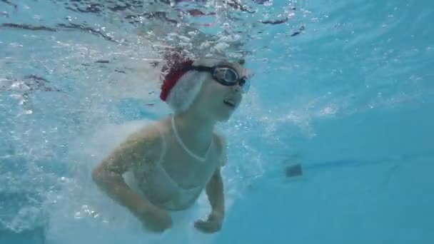 Милая маленькая девочка в красной шляпе Санты занимается спортом в детском бассейне. Она плавает под водой в пузырьках, смотрит в камеру и улыбается. Рождество. Концепция. Портрет. Закрывай. Медленное движение. — стоковое видео