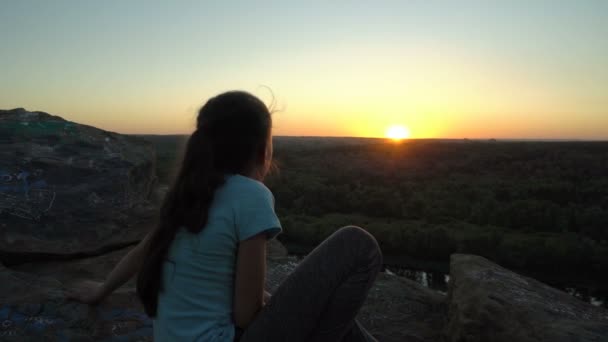 Menina feliz olha para o pôr-do-sol, sentado no topo de uma montanha sobre um rio e um vale verde. Conceito. Close-up. Visão traseira. 4K. — Vídeo de Stock