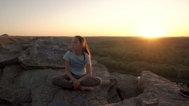 A sweet, brooding teenage girl sits on top of a cliff against the yellow setting sun. Her hair flutters in the wind. Beautiful landscape at sunset. Portrait. 4K. — Stock Video