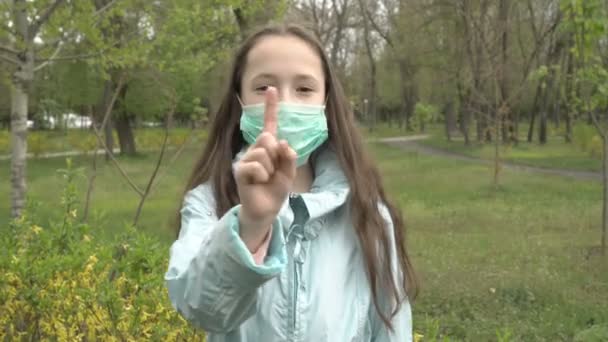 Een schattig klein meisje met een medisch masker schudt met haar vinger waarschuwend voor de camera terwijl ze in een leeg stadspark staat. Portret. Concept. Coronavirus. Tweede golf. Preventie. Een close-up. 4K. — Stockvideo