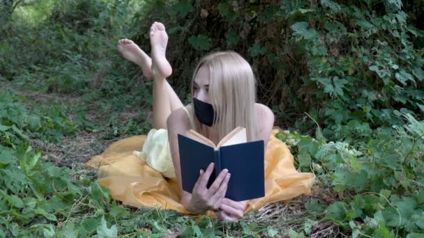 Une belle blonde avec un masque noir sur le visage pose pour une photographe, allongée dans la nature sur l'herbe avec un livre bleu à la main. Ferme là. Portrait. 4K. — Video