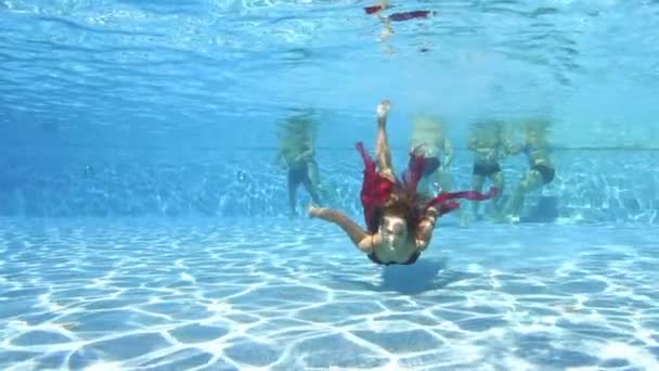 Menina adolescente bonito nada em um vestido vermelho na câmera subaquática em uma piscina pública ao ar livre. Ela sorri e acena as mãos. Uma rapariga com um cabelo comprido bonito. Retrato. Conceito. Movimento lento — Vídeo de Stock