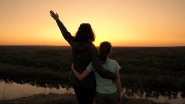 Anne ve küçük kızı gün batımında tepenin üstünde durur, sarılır ve güneşe el sallarlar. Siluetler. Dikiz aynası. Kavram. 4K