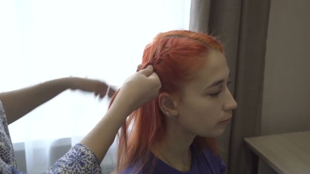 大人の女性は、早朝の部屋の明るい窓を背景に、赤い髪をした10代の女の子の頭の上に編組します。肖像画。閉めろ。背景をぼんやり。4K — ストック動画