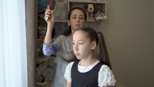 A mother combs her daughters hair in her school uniform and ties her tails in the early morning in front of her room window. Mom is getting her daughter ready for school. Portrait. Close-up. 4K — Stock Video