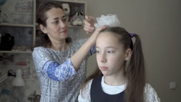 Seorang wanita Oriental menghiasi kepala seorang gadis kecil Eropa dengan busur putih di sebuah ruangan di pagi hari. Konsep hubungan internasional. Potret. Close-up. 4K — Stok Video