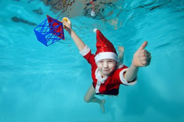 Suyun altında Noel Baba kostümü giymiş neşeli küçük bir çocuğun yakın çekimi. Yeni yıl hediyesiyle havuzun dibine daldı ve baş parmağını kaldırdı. Aktif mutlu çocuk. Sağlıklı yaşam tarzı