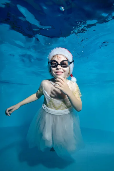 Retrato de una niña sonriente con un vestido blanco en el fondo de una piscina infantil con un juguete de Navidad. Decoraciones en forma de árboles de Navidad en la cabeza. Clase de natación —  Fotos de Stock