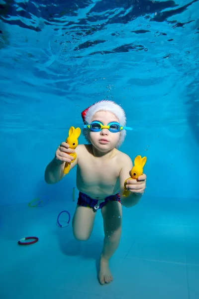Retrato de un niño en un sombrero de Santa Claus y gafas en una piscina bajo el agua. El niño aprende a divertirse buceando bajo el agua. Tiene juguetes en sus manos. Clases de natación con un niño. Estilo de vida saludable — Foto de Stock