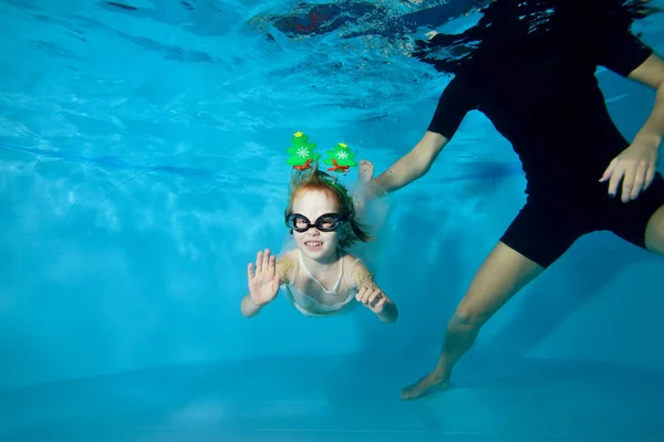 Una niña con un vestido está nadando bajo el agua en la piscina. Decoraciones navideñas en la cabeza. Clases con el bebé. Clase de natación. Estilo de vida saludable. Orientación horizontal — Foto de Stock