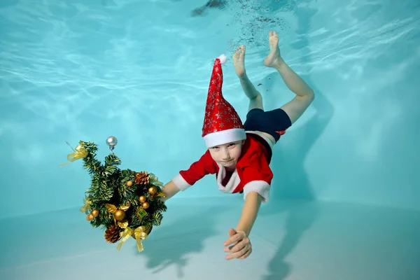 Portret chłopca pod wodą w kostiumie Świętego Mikołaja w basenie. Trzyma choinkę. Aktywność na zewnątrz. Zdrowy styl życia. Szkoła pływania. Koncepcja uroczystości — Zdjęcie stockowe