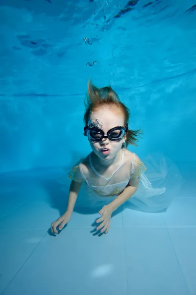 Hermosa niña bajo el agua en el fondo de la piscina. Ella sonríe y mira a la cámara. El bebé aprende a bucear. Clases de natación con un niño. Ejercicios corporales. Orientación vertical —  Fotos de Stock