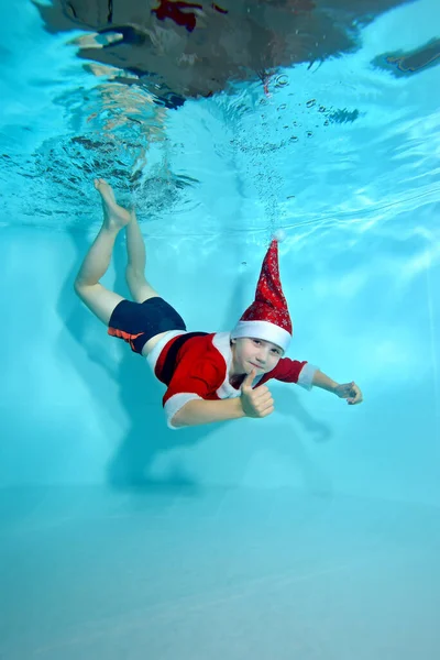 Funny little boy in Santa Claus costume underwater. He dives to the bottom of the pool and gives a thumbs up. Active child. Healthy lifestyle. Swimming lessons underwater