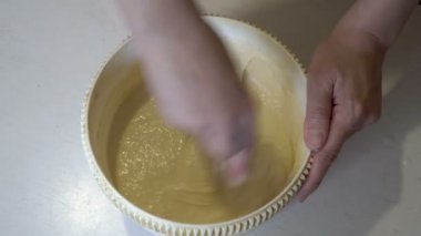 Bir kadın evin mutfağındaki yuvarlak bir kasede hamuru beyaz ahşap bir masada çırpıyor. Ev yapımı yemek pişirme kavramı. Yakın plan. Yukarıdan manzara. Yüzsüz. 4K