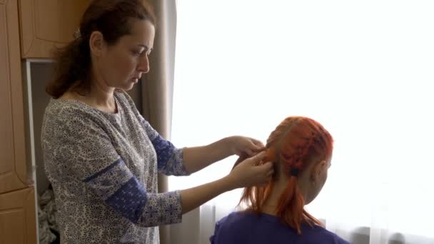 東洋の女性は、太陽の光と窓の近くの部屋の椅子に彼女の前に座っている赤-髪の10代の少女の髪を編みます。ヘアケアの概念。接近中だ。4K — ストック動画