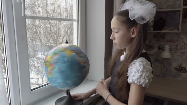 Una bambina triste sta alla finestra con un globo in mano e fiocchi bianchi sulla testa. Gira il globo velocemente e guarda pensieroso fuori dalla finestra. Il concetto di auto-isolamento. 4K — Video Stock