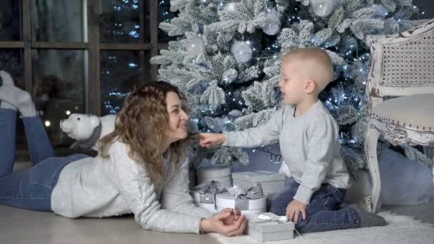 おもちゃで美しいクリスマスツリーの前に横たわる幸せなお母さんと赤ちゃんの遊び。彼らは笑い、周りをばかにし、プレゼントを見てください。幸せな新年の概念。接近中だ。4K — ストック動画
