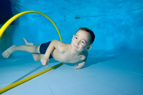 Un hermoso niño está nadando bajo el agua en una piscina para niños. Nada a través del aro, saca juguetes. Niño feliz activo. Estilo de vida saludable. Clases de natación bajo el agua. Un deporte familiar — Foto de Stock
