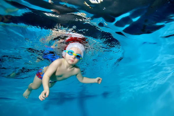 Retrato de un niño en un sombrero de Navidad y vasos en una piscina bajo el agua. Mira a la cámara. El bebé aprende a bucear. Clases de natación con un niño. Estilo de vida saludable. Orientación horizontal — Foto de Stock