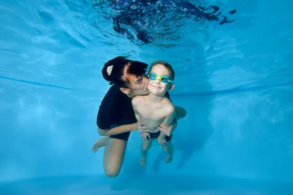 Un hermoso niño y su madre nadan juntos bajo el agua. Ella lo besa suavemente en la mejilla. Niño feliz activo. Clases de deportes familiares. Estilo de vida saludable. Clase de natación bajo el agua — Foto de Stock