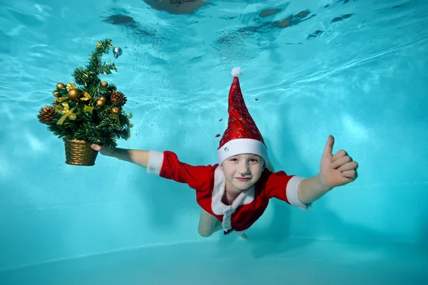 산타 클로스 복장을 한 쾌활 한 소년의 크리스마스 트리가 있는 수중 사진. 그는 수영장 바닥으로 잠수하여 박수를 친다. 활동적 인아이. 건강 한 생활 방식 — 스톡 사진