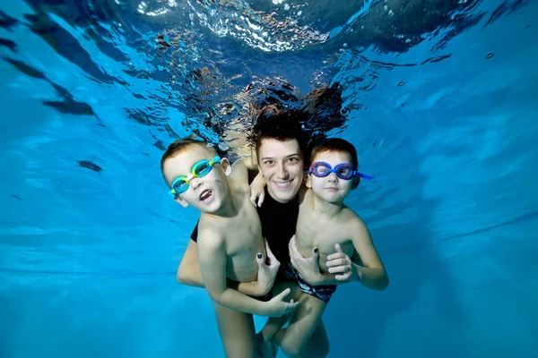 Dos hermosos chicos bajo el agua con su madre. Los abraza tiernamente. Sonríen y miran a la cámara. Niños felices activos. Estilo de vida saludable. Clases de natación bajo el agua. Deportes familiares — Foto de Stock