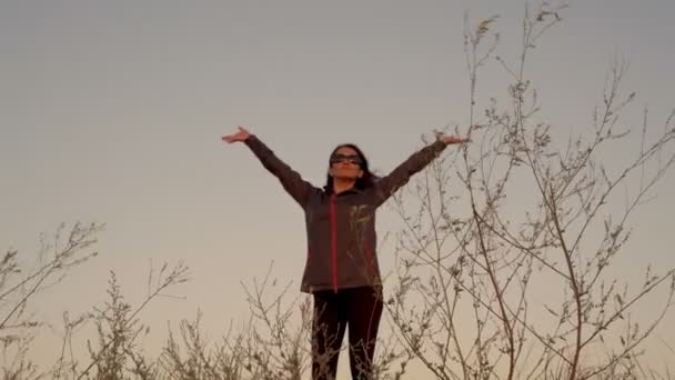 Eine glückliche Frau mittleren Alters meditiert und streckt sich im Sonnenuntergang in Sportkleidung an einem Sommerabend auf einem Berggipfel stehend. Der Fokus verlagert sich vom Gras im Vordergrund auf die Frau. 4K — Stockvideo