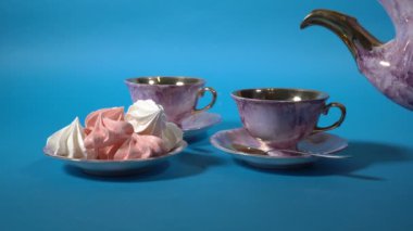 Yakından, bir kadın yavaşça pembe çaydanlıktan güzel yaldızlı porselen bardaklara çay dolduruyor. Pembe fincanlar mavi bir masanın üzerinde duruyor. Durgun yaşam. Yüzsüz. Konukseverlik kavramı. 4K