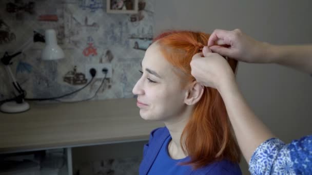 Uma adolescente doce e sorridente está sentada em um quarto perto da janela, e sua mãe está trançando seu cabelo vermelho em tranças. O conceito de mães se preocupa. Retrato de uma mulher. Vista lateral. Close-up. 4K — Vídeo de Stock