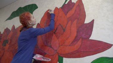 Kızıl saçlı güzel bir genç kız, salgın nedeniyle karantina sırasında fırçayla duvarına büyük bir kırmızı çiçek çizer. Konsept. Yakın plan. Alt manzara. 4K